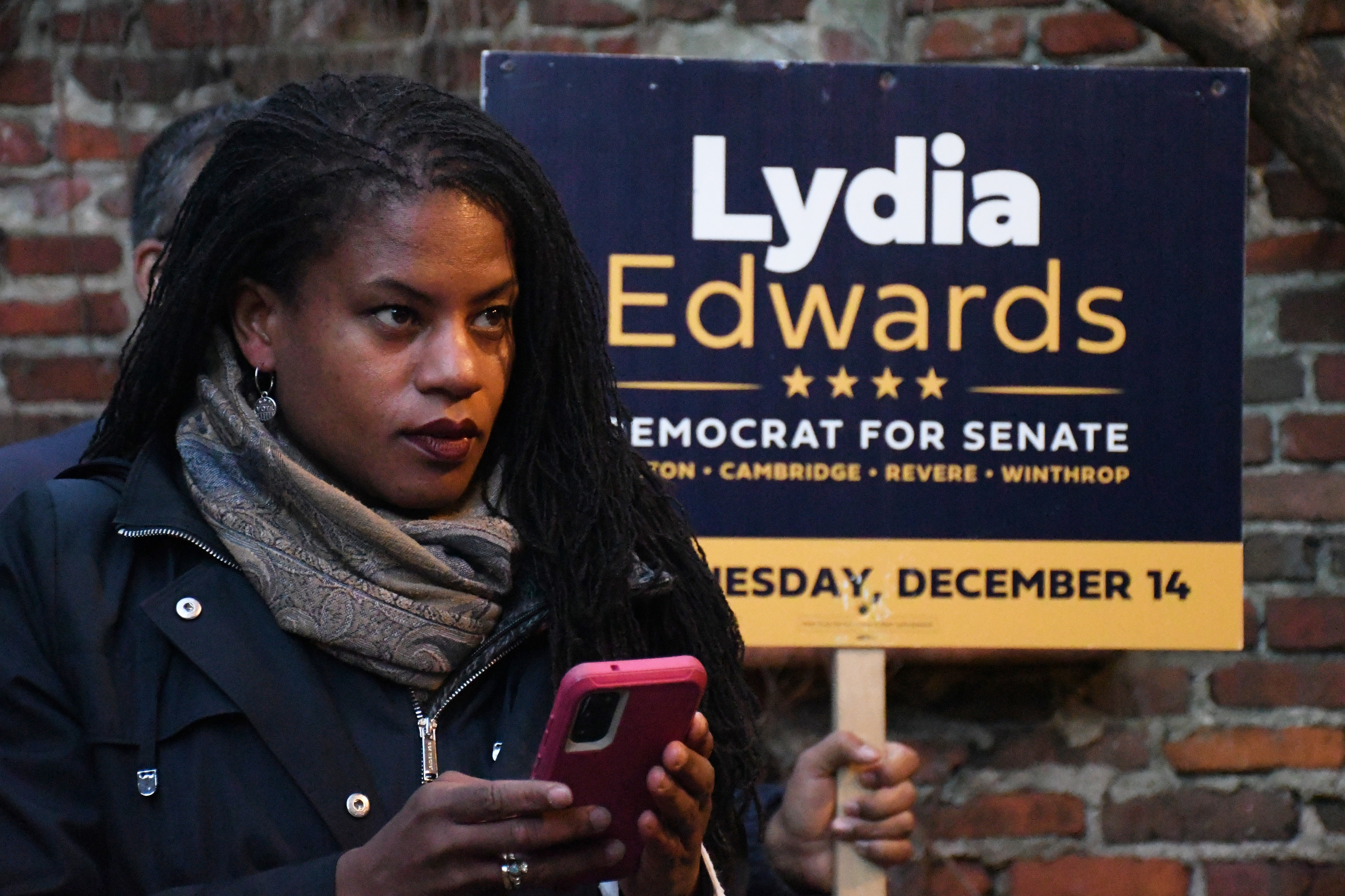 A Black woman uses her phone. A sign behind her says 'Lydia Edwards: Democrat for Senate'.