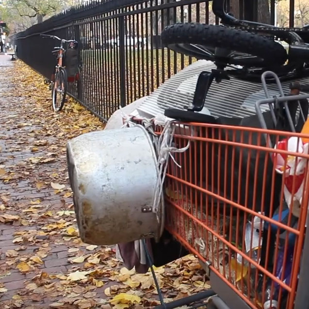 A shopping cart filled with someone's belongings sits next to a fence.