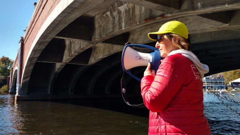 a woman in a red jacket stands with a megaphone on a riverbank. There is a large brick bridge behind her.